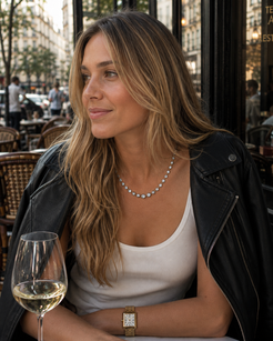 A woman with long blonde-highlighted hair sits at an outdoor cafe table wearing a silver beaded necklace, a white tank top, a black leather jacket, and a gold watch. A glass of white wine is in the foreground.